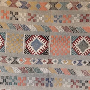May include: A detailed close-up of a woven textile with a light gray background. The design features repeating geometric patterns in various colors, including red, blue, orange, and yellow. The textile appears to be a rug or fabric with a textured surface.