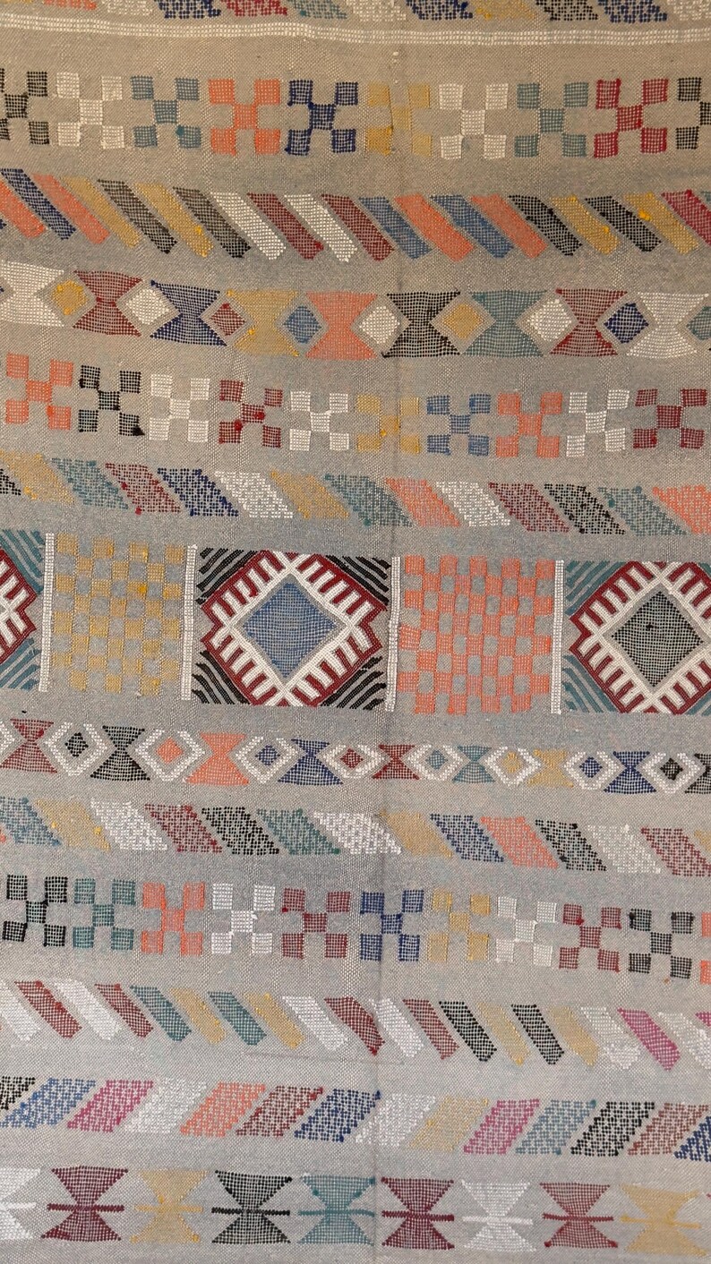 May include: A detailed close-up of a woven textile with a light gray background. The design features repeating geometric patterns in various colors, including red, blue, orange, and yellow. The textile appears to be a rug or fabric with a textured surface.