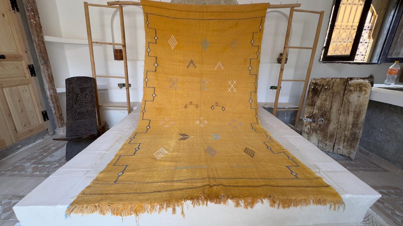 Pu&ograve; includere: Tappeto giallo senape con motivi geometrici e frange. Il tappeto presenta disegni a rombi, frecce e zig-zag in bianco e nero. Il tappeto &egrave; esposto su una superficie bianca, con un telaio in legno sullo sfondo.