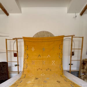 Pu&ograve; includere: Tappeto giallo senape con motivi geometrici e frange. Il tappeto presenta disegni a rombi, a zig-zag e a croce in bianco, nero e blu. Il tappeto &egrave; esposto su una cornice di legno, che ne mette in risalto la trama e il colore.