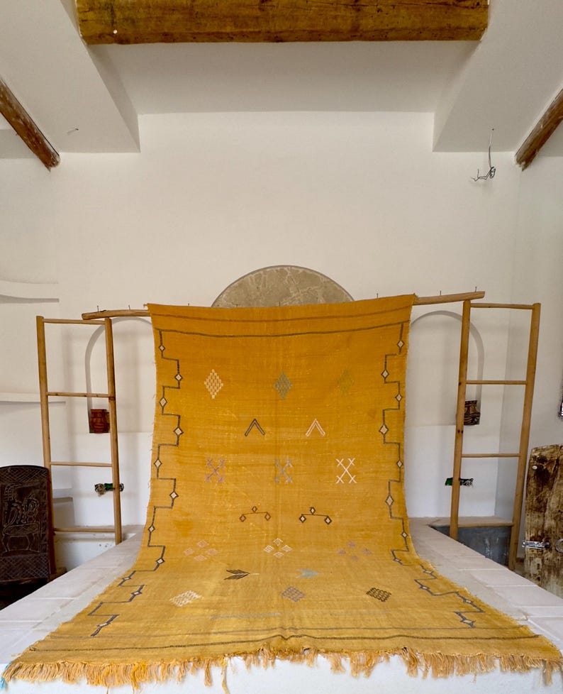 Pu&ograve; includere: Tappeto giallo senape con motivi geometrici e frange. Il tappeto presenta disegni a rombi, a zig-zag e a croce in bianco, nero e blu. Il tappeto &egrave; esposto su una cornice di legno, che ne mette in risalto la trama e il colore.
