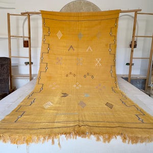 Pu&ograve; includere: Tappeto giallo senape con motivi geometrici e frange. Il tappeto presenta disegni a forma di diamante, triangolo e croce in vari colori. Il tappeto &egrave; esposto su una superficie bianca, mettendo in risalto i suoi intricati dettagli e la sua consistenza.