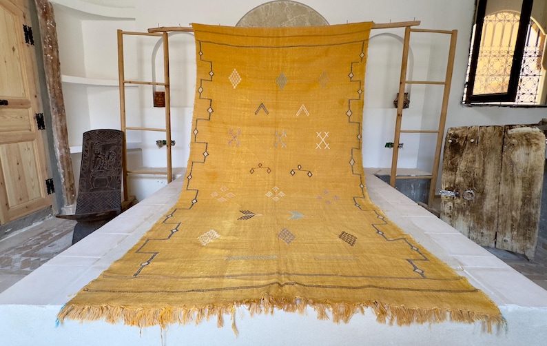 Pu&ograve; includere: Tappeto giallo senape con motivi geometrici e frange. Il tappeto presenta disegni a forma di diamante, triangolo e croce in vari colori. Il tappeto &egrave; esposto su una superficie bianca, mettendo in risalto i suoi intricati dettagli e la sua consistenza.