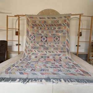 May include: A large, rectangular woven rug with a geometric pattern in beige, blue, pink, and orange. The rug has a fringe along the bottom edge and is displayed on a white surface. The rug is a decorative home decor item.