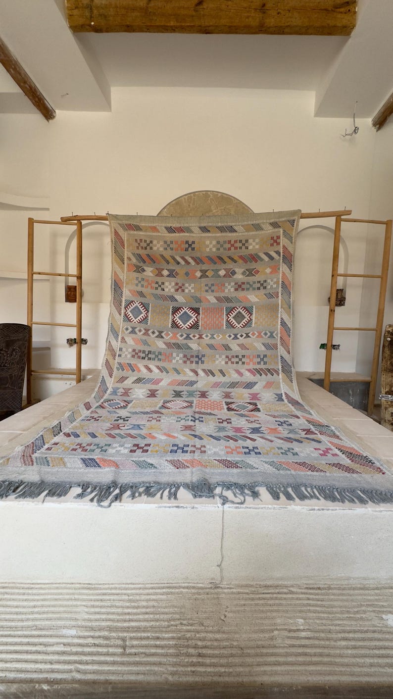 May include: A large, rectangular woven rug with a geometric pattern in beige, blue, pink, and orange. The rug has a fringe along the bottom edge and is displayed on a white surface. The rug is a decorative home decor item.