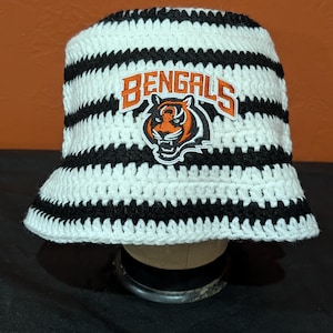 May include: A crocheted bucket hat featuring horizontal black and white stripes. The front of the hat displays an embroidered patch with the word BENGALS in orange letters above a graphic of a tiger head in orange, black, and white colours.
