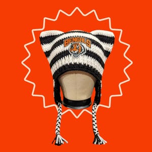 Peut inclure: Un bonnet en crochet à rayures noires et blanches avec des oreilles de chat, arborant le logo des Cincinnati Bengals avec une tête de tigre et le texte BENGALS. Le bonnet comporte deux longues tresses suspendues sur les côtés en laine texturée.