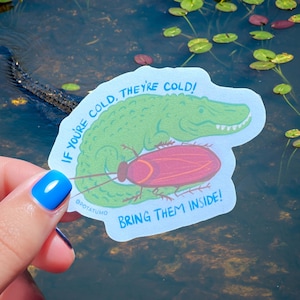 May include: A sticker featuring a green cartoon alligator and a pink cartoon cockroach. The sticker has the text "IF YOU'RE COLD, THEY'RE COLD! BRING THEM INSIDE!" The sticker is held in front of a body of water.