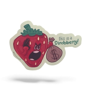 May include: A die-cut sticker featuring a cartoon strawberry with an angry expression, holding a money bag. The text "this is a Strobberry!" is written on the sticker. The sticker has a white border and a shadow.