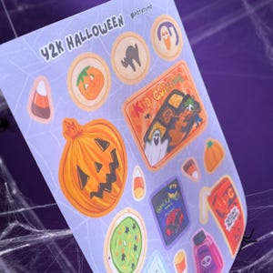 May include: A sheet of stickers with a Y2K Halloween theme. The stickers feature various Halloween-related designs, including pumpkins, candy corn, ghosts, and the text "Kid Crazing". The color palette includes orange, purple, and green.