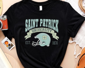 May include: Black t-shirt with a white and green graphic design that says "Saint Patrick University Est. 1831". The graphic includes a green shamrock and a football helmet.