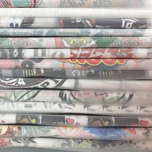May include: A stack of folded t-shirts with various colorful designs and graphics. The shirts feature different characters, logos, and text. The shirts are wrapped in clear plastic.