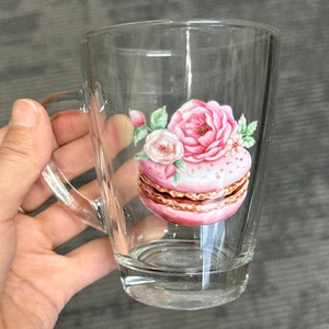May include: A clear glass mug with a handle features a pink and white macaron with floral accents. The macaron is decorated with white dots and a brown layer.