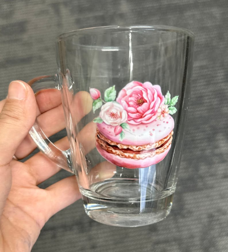 May include: A clear glass mug with a handle features a pink and white macaron with floral accents. The macaron is decorated with white dots and a brown layer.