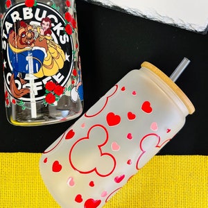 May include: Two glass tumblers with lids and straws. The first tumbler has a frosted finish with red hearts and Mickey Mouse ears. The second tumbler has a clear glass with a printed image of Belle and the Beast from Disney's Beauty and the Beast. The image also includes the Starbucks logo and the text "Starbucks Coffee".