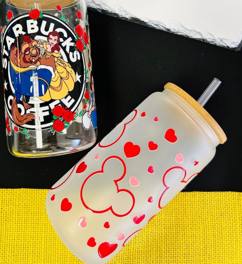 May include: Two glass tumblers with lids and straws. The first tumbler has a frosted finish with red hearts and Mickey Mouse ears. The second tumbler has a clear glass with a printed image of Belle and the Beast from Disney's Beauty and the Beast. The image also includes the Starbucks logo and the text "Starbucks Coffee".