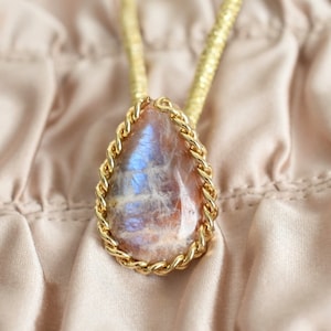 May include: A gold chain necklace with a teardrop-shaped iridescent gemstone pendant. The pendant is set in a gold rope-like frame.