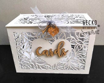 May include: White wooden box with a heart-shaped cutout and the word "cards" on it. The box has a delicate floral design and a key with a heart-shaped charm attached to it. The box is for holding wedding cards.