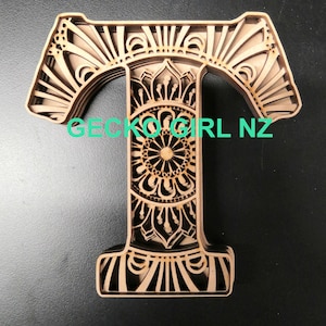 May include: A wooden letter T with a mandala design. The letter is intricately cut and layered, creating a 3D effect. The letter is painted in a light brown color.