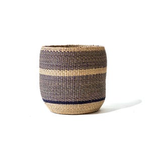 Op de afbeelding: Een cilindrische geweven mand met een natuurlijke beige basis en donkerblauwe horizontale strepen. De mand is gemaakt van strak geweven vezels en heeft een stevige, afgeronde vorm. De mand is geschikt voor opslag of decoratie.