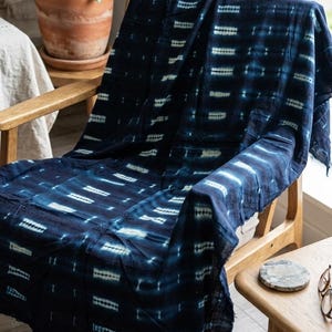 May include: A dark blue textile throw blanket featuring a repeating pattern of horizontal white tie-dye stripes. The fabric has a visible weave and frayed edges, draped over a wooden chair. The pattern consists of small, dashed white marks.