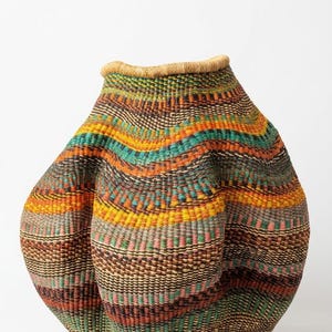 Può includere: Un cesto intrecciato a mano con una forma bulbosa e asimmetrica e un'ampia apertura. L'esterno presenta fasce orizzontali di colori vivaci tra cui arancione, giallo, verde acqua, rosa e marrone scuro, con bordo e base intrecciati color naturale.