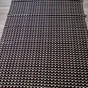 Peut inclure: Un tapis rectangulaire tissé avec un motif répétitif de carrés noirs, marron et blancs. Le tapis a une bordure beige et est posé sur un sol en bois clair. Le motif crée un aspect texturé.