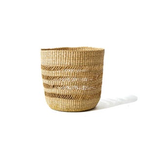 Puede incluir: Una cesta cilíndrica tejida, de color marrón claro natural. La cesta presenta bandas horizontales de tejido abierto, creando un patrón texturizado. La cesta se utiliza probablemente para almacenamiento o decoración.