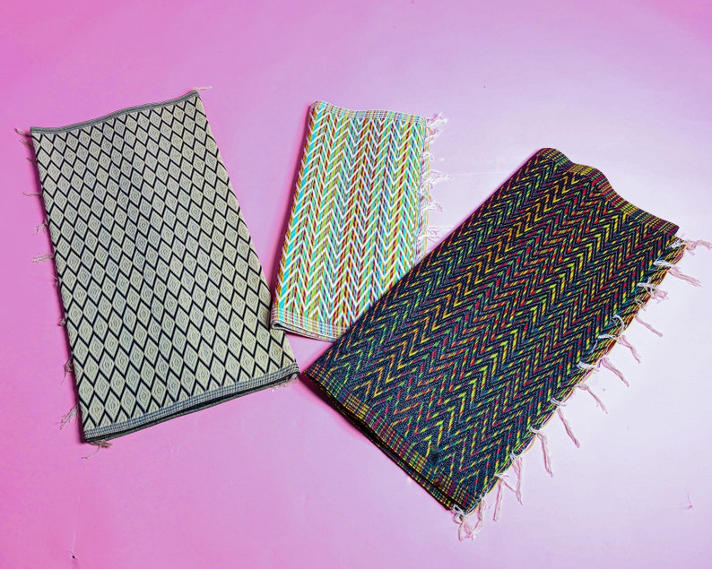 May include: Three woven mats with different patterns. The first mat is beige with a black diamond pattern. The second mat is white with a multi-colored chevron pattern. The third mat is black with a multi-colored chevron pattern.
