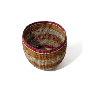Peut inclure: Un panier rond tissé à la main, avec une large ouverture. Il présente un motif rayé dans des tons de marron, beige et gris, avec un bord magenta. Les matériaux naturels et les tons terreux suggèrent une esthétique rustique et artisanale. Le diamètre du panier est d'environ 20 cm.