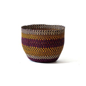 Puede incluir: Una cesta tejida con forma redondeada. La cesta presenta un llamativo patrón de bandas alternas en amarillo, burdeos y azul oscuro. Está hecha de fibras naturales y tiene una superficie texturizada.