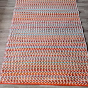 Peut inclure: Un tapis rectangulaire avec un motif en chevrons vibrant dans les tons orange, rouge, blanc et bleu. Le tapis a une bordure orange unie et est présenté sur un sol en bois. Le motif en zigzag crée un aspect texturé.
