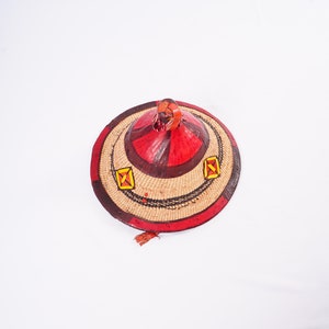 Peut inclure: Un chapeau traditionnel en paille tressée de forme conique, orné de motifs géométriques rouges, bruns et jaunes. Le chapeau a un petit pompon qui pend en bas.