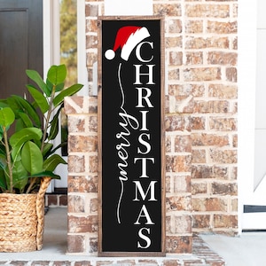 Puede incluir: Un letrero de madera negro con letras blancas que dice "feliz navidad" con un gorro de Papá Noel rojo en la parte superior.