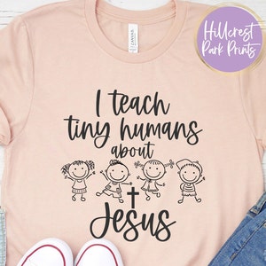 May include: A pink t-shirt with the text "I teach tiny humans about Jesus" and a graphic of four children holding hands around a cross.