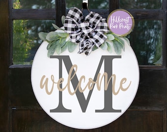 Op de afbeelding: Ronde witte houten deurhanger met een zwart-wit geblokte strik, groene bladeren en de letter "M" in grijs met het woord "welkom" in beige schrift.