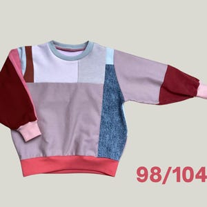 Könnte beinhalten: Ein Patchwork-Sweatshirt in verschiedenen Farben, darunter Rosa, Burgunderrot und Blau. Die langen Ärmel haben eine Ballonform und die Bündchen sind rosa. Der untere Saum ist korallenfarben und der Ausschnitt hellblau. Die Zahl "98/104" ist unten rechts aufgedruckt.