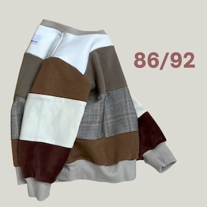Könnte beinhalten: Ein braunes, weißes und graues Patchwork-Sweatshirt mit braunem Samtkragen und -bündchen. Das Sweatshirt hat ein weißes Etikett mit dem Text "86/92" auf der Vorderseite.
