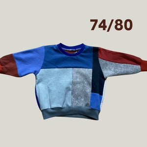 Könnte beinhalten: Ein Sweatshirt im Patchwork-Stil in verschiedenen Farben, darunter Blau, Rot, Grau und Schwarz. Das langärmlige Oberteil hat einen gerippten Ausschnitt und Bündchen. Die Größe 74/80 ist braun rechts oben aufgedruckt.