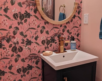 May include: A white porcelain bathroom sink with a gold faucet and a black vanity cabinet. The sink is set against a pink wall with a floral wallpaper featuring a leopard print design.