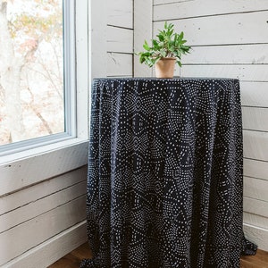 May include: A black and white patterned tablecloth with a small potted plant on top. The tablecloth is draped over a round table and the table is in a room with white wood paneling.