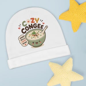 May include: A white baby beanie featuring a colorful graphic of a smiling bowl of congee with a spoon. The text reads COZY CONGEE and WARM BELLY, HAPPY HEART. The design includes small red hearts and green accents, presented on a plain white fabric.
