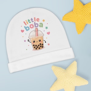 Pode incluir: Um gorro branco para bebé com um gráfico colorido de um copo de chá boba sorridente com pérolas de tapioca pretas, uma palhinha cor-de-rosa e o texto "little boba" em letras multicoloridas, rodeado por pequenos corações e estrelas em tons pastel.
