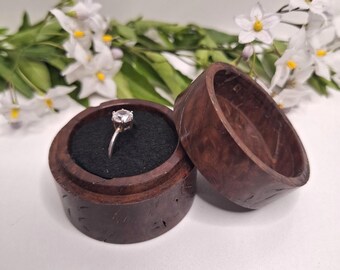May include: A silver engagement ring with a clear gemstone sits in an open, round, dark brown wooden ring box with a black velvet interior. The box is next to its lid. White flowers with yellow centers and green leaves are in the background.