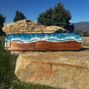May include: A rectangular wooden serving tray with a resin ocean design. The tray features a dark wood base with a blue and white resin wave pattern. It has a metal handle and is set on a large rock.