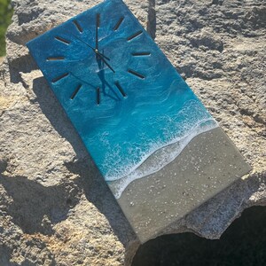 May include: Rectangular wall clock with a beach-themed design. The clock face features a gradient of blue and white, resembling ocean waves, with a sandy-colored bottom. Black clock hands and hour markers complete the design. The clock is displayed on a large rock.