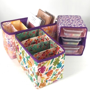 Puede incluir: Tres cajas de almacenamiento de tela con estampados florales y de hojas. Las cajas tienen ribetes y separadores morados. Una caja contiene libros, otra recipientes transparentes y la tercera está vacía.