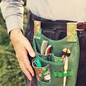 Op de afbeelding: Een groene tuingereedschapsriem met meerdere zakken en een voering met bloemenprint. De riem wordt om de taille gedragen en heeft een bruine leren riem met een zilveren gesp.