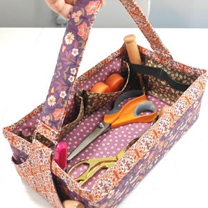 May include: A patterned fabric craft organiser with a handle, in shades of purple, orange, and white. It contains sewing tools such as scissors, wooden tools, and orange spools. The interior fabric has a polka dot design.