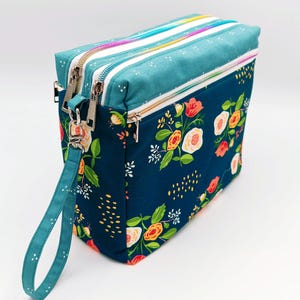 May include: A teal and navy blue zippered pouch with a floral print and a teal strap. The pouch has three zippered compartments.